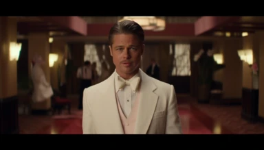 a screengrab of Brad Pitt as Aldo Raine from the movie Inglourious Basters in the style of the director Wes Anderson, full body shot, he is in the center of the photograph looking directly at the camera. He has a pencil moustache. He is dressed in a white tuxedo jacket, white shirt, black bow-tie, red rose lapel clip, black pants. The background is an art decó movie theatre designed in the style of Wes Anderson in a Wes Anderson color palette, symmetrical, whimsical, ornate, planimetric composit